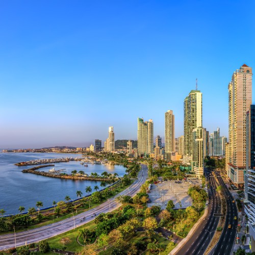 Skyline of a coastal city under a clear blue sky, with tall modern skyscrapers, a bustling road, and a serene waterfront lined with palm trees.