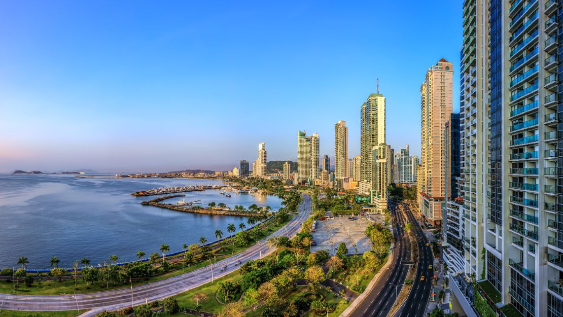 Skyline of a coastal city under a clear blue sky, with tall modern skyscrapers, a bustling road, and a serene waterfront lined with palm trees.
