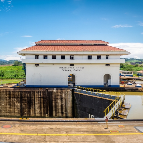 Panama Canal's Miraflores Locks on a sunny day, featuring the control building with a red-tiled roof, surrounded by greenery and blue skies.