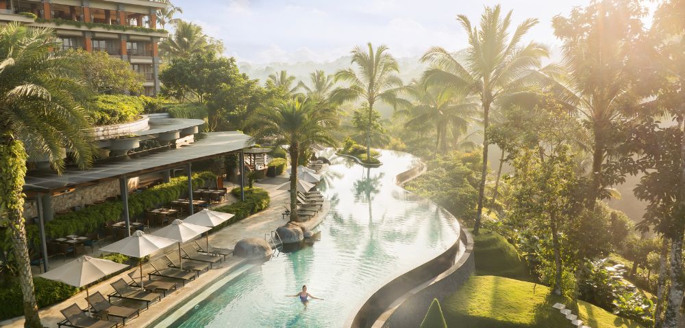 A serene infinity pool at a tropical resort, surrounded by palm trees and sun loungers, with a person swimming. Soft sunlight creates a tranquil ambiance.