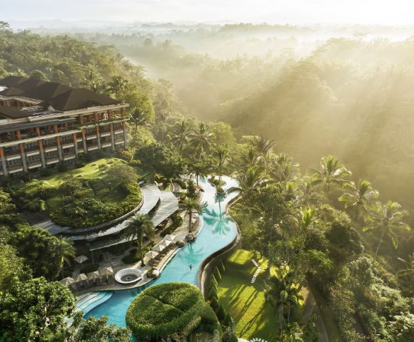 Aerial view of a luxury resort surrounded by lush greenery. A winding pool with sun loungers and palm trees under scattered sunlight creates a serene atmosphere.