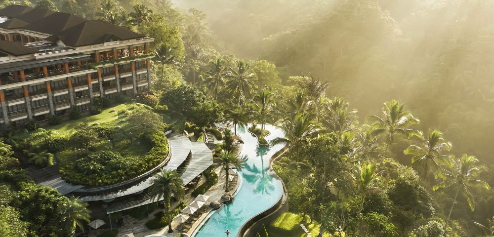 Aerial view of a luxury resort surrounded by lush greenery. A winding pool with sun loungers and palm trees under scattered sunlight creates a serene atmosphere.