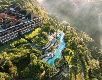 Aerial view of a lush resort with a winding pool surrounded by vibrant greenery and tropical trees. A large building is on the left, with mist over the jungle. Luxurious and tranquil atmosphere.
