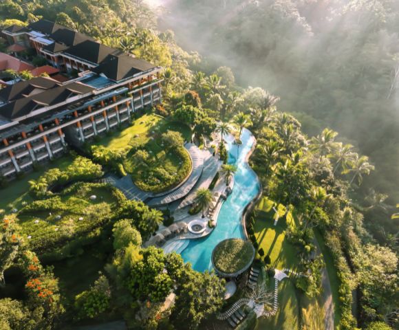Aerial view of a lush resort with a winding pool surrounded by vibrant greenery and tropical trees. A large building is on the left, with mist over the jungle. Luxurious and tranquil atmosphere.