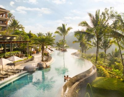 Luxurious resort with an infinity pool surrounded by palm trees and greenery. A couple relaxes in the pool, exuding a calm, serene atmosphere.