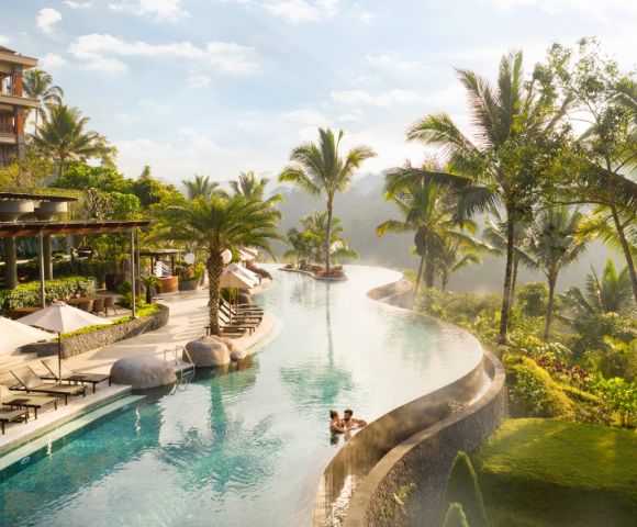 Luxurious resort with an infinity pool surrounded by palm trees and greenery. A couple relaxes in the pool, exuding a calm, serene atmosphere.