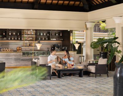 A couple sits comfortably in a sophisticated lounge with tropical plants and a stocked bar in the background, creating a relaxed and elegant atmosphere.