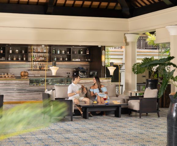 A couple sits comfortably in a sophisticated lounge with tropical plants and a stocked bar in the background, creating a relaxed and elegant atmosphere.