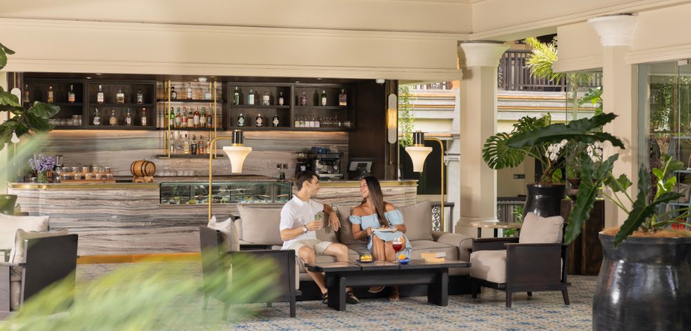 A couple sits comfortably in a sophisticated lounge with tropical plants and a stocked bar in the background, creating a relaxed and elegant atmosphere.