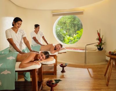 Two women receive relaxing massages on tables in a serene spa. The room, with wooden floors and a circular window revealing lush greenery, conveys tranquility.
