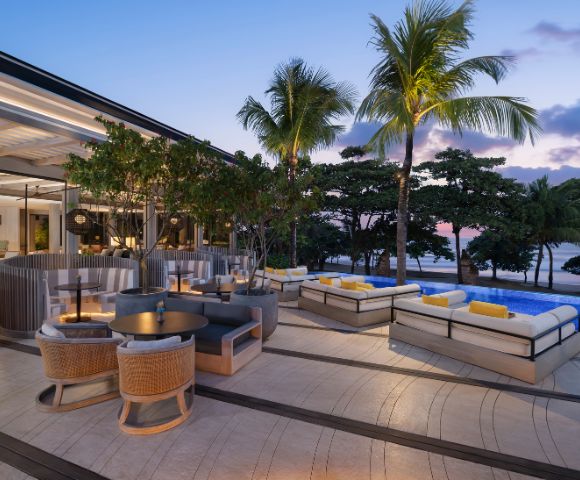 Elegant beachside lounge at sunset, featuring modern wicker furniture, plush cushions, and ambient lighting. Palm trees frame the serene ocean view.
