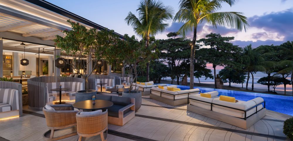 Elegant beachside lounge at sunset, featuring modern wicker furniture, plush cushions, and ambient lighting. Palm trees frame the serene ocean view.