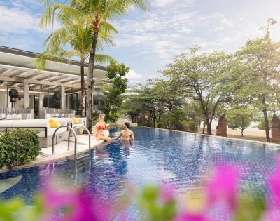 A couple relaxes in a serene hotel pool surrounded by lush greenery and a beach view. The scene is bright and tranquil, with tropical vibes.