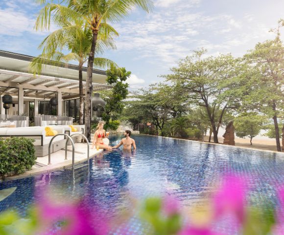 A couple relaxes in a serene hotel pool surrounded by lush greenery and a beach view. The scene is bright and tranquil, with tropical vibes.