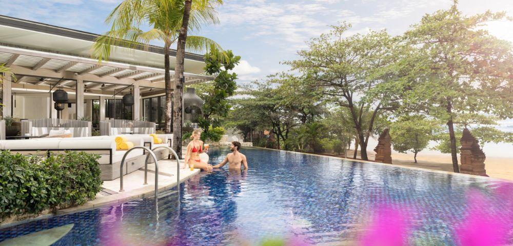A couple relaxes in a serene hotel pool surrounded by lush greenery and a beach view. The scene is bright and tranquil, with tropical vibes.