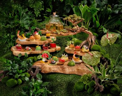 A whimsical woodland-themed tea display features intricate pastries and colorful treats on tiered wooden platters, surrounded by lush greenery and moss.