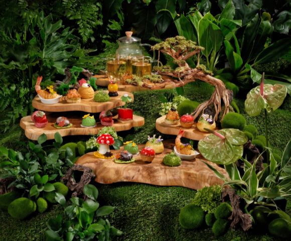 A whimsical woodland-themed tea display features intricate pastries and colorful treats on tiered wooden platters, surrounded by lush greenery and moss.