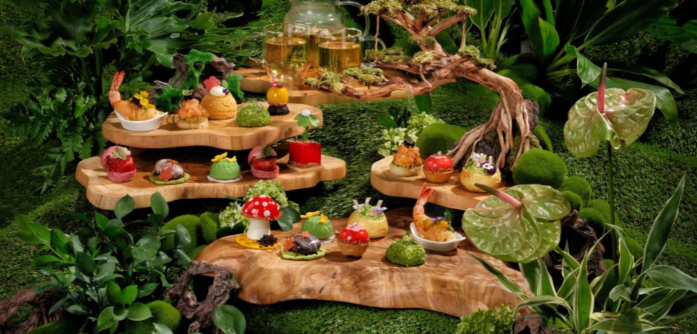 A whimsical woodland-themed tea display features intricate pastries and colorful treats on tiered wooden platters, surrounded by lush greenery and moss.