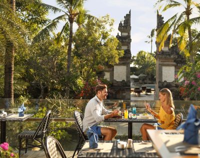 A couple dines outdoors at a tropical restaurant, surrounded by lush greenery and palm trees. The setting is tranquil, evoking a romantic atmosphere.