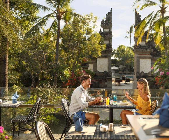 A couple dines outdoors at a tropical restaurant, surrounded by lush greenery and palm trees. The setting is tranquil, evoking a romantic atmosphere.