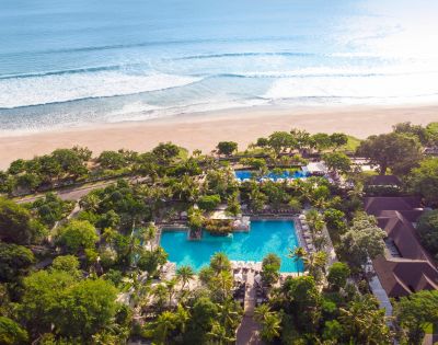 Aerial view of a tranquil beach resort with multiple pools surrounded by lush greenery, leading to a sandy beach and gentle ocean waves in the background.