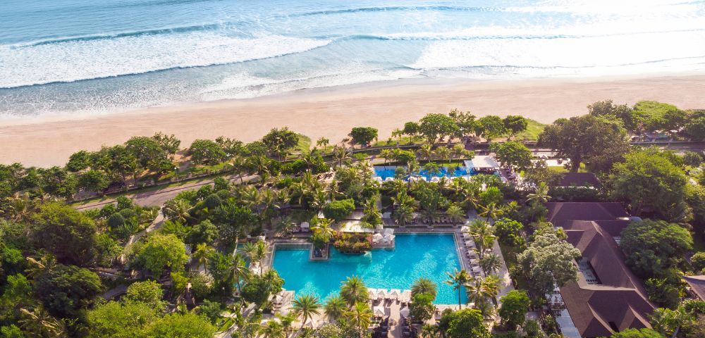 Aerial view of a tranquil beach resort with multiple pools surrounded by lush greenery, leading to a sandy beach and gentle ocean waves in the background.