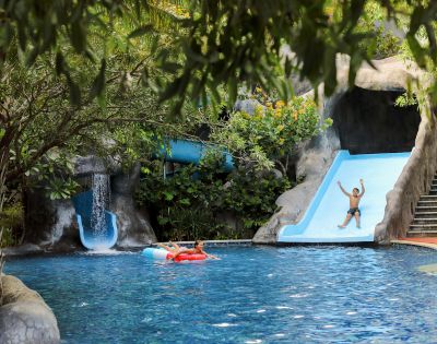 A vibrant pool scene with lush greenery, featuring a person joyfully sliding down a blue waterslide. Another person relaxes on a red float in the water.