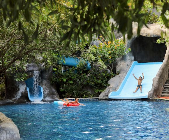 A vibrant pool scene with lush greenery, featuring a person joyfully sliding down a blue waterslide. Another person relaxes on a red float in the water.