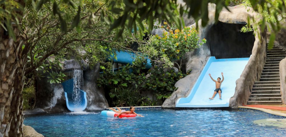 A vibrant pool scene with lush greenery, featuring a person joyfully sliding down a blue waterslide. Another person relaxes on a red float in the water.