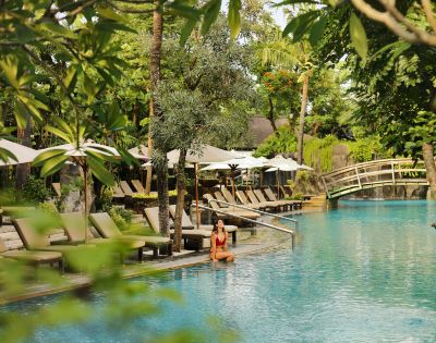 A woman in a red swimsuit sits by a tranquil pool surrounded by lush greenery. Beige lounge chairs and white umbrellas line the poolside, with a wooden bridge in the background, evoking a serene, tropical atmosphere.