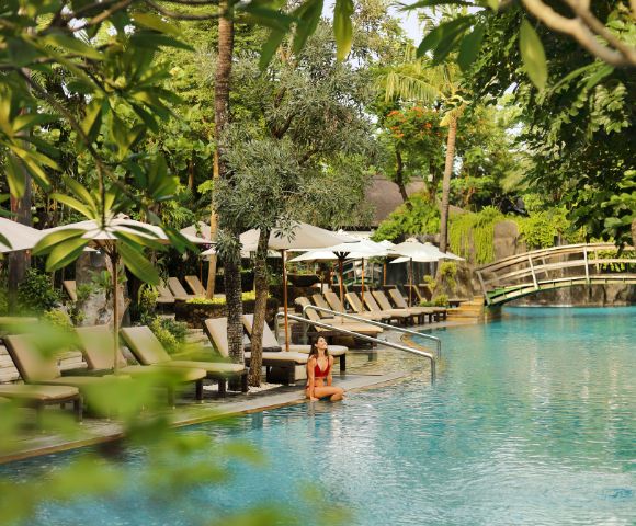 A woman in a red swimsuit sits by a tranquil pool surrounded by lush greenery. Beige lounge chairs and white umbrellas line the poolside, with a wooden bridge in the background, evoking a serene, tropical atmosphere.