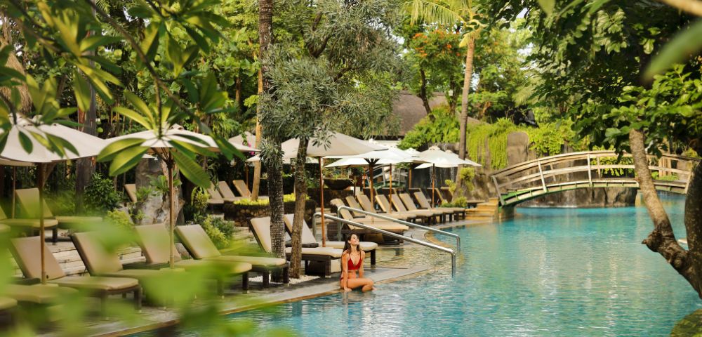 A woman in a red swimsuit sits by a tranquil pool surrounded by lush greenery. Beige lounge chairs and white umbrellas line the poolside, with a wooden bridge in the background, evoking a serene, tropical atmosphere.