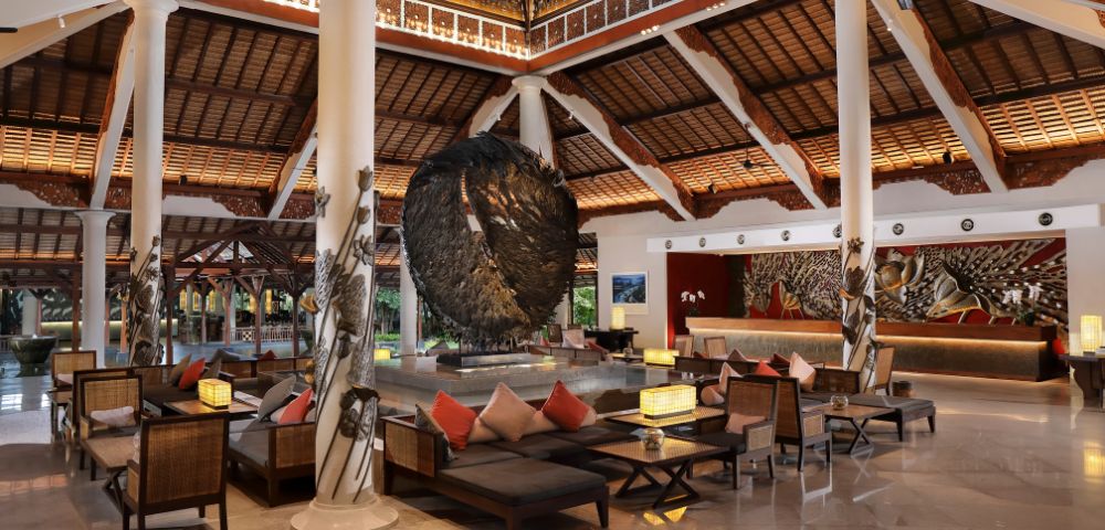 Spacious, elegant hotel lobby with high wooden ceilings, large circular sculpture, and comfortable seating. Warm lighting creates a welcoming atmosphere.