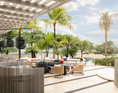 Outdoor beachside dining area with modern seating and palm trees. A couple enjoys a meal under a pergola, overlooking sand and sea. Relaxed ambiance.