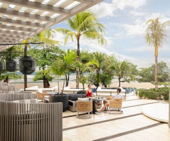 Outdoor beachside dining area with modern seating and palm trees. A couple enjoys a meal under a pergola, overlooking sand and sea. Relaxed ambiance.