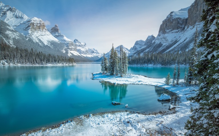 Snow-covered mountains and pine trees surround a tranquil turquoise lake under a clear blue sky, creating a serene and peaceful winter scene.