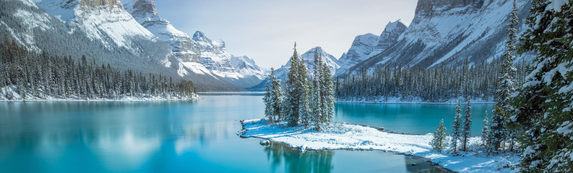 Snow-covered mountains and pine trees surround a tranquil turquoise lake under a clear blue sky, creating a serene and peaceful winter scene.
