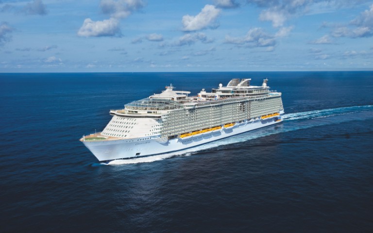 A large white cruise ship sails through a vast, calm ocean under a clear blue sky. The ship features multiple decks and geometric structures.