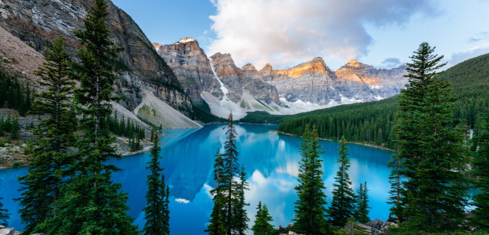 A serene mountain lake scene with calm turquoise waters reflecting surrounding pine trees and rugged peaks under a partly cloudy sky, evoking tranquility.