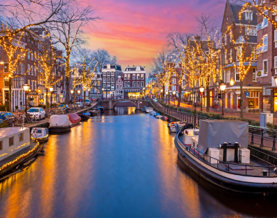 A scenic Amsterdam canal at sunset, adorned with fairy lights along the trees and buildings, reflecting in the water.