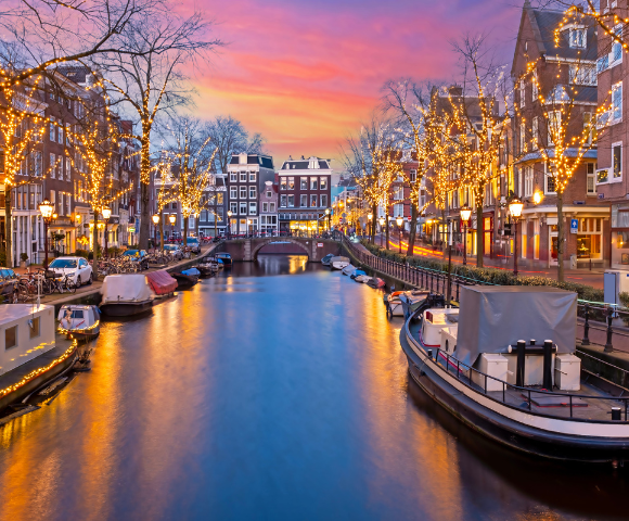A scenic Amsterdam canal at sunset, adorned with fairy lights along the trees and buildings, reflecting in the water.