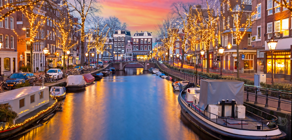 A scenic Amsterdam canal at sunset, adorned with fairy lights along the trees and buildings, reflecting in the water.