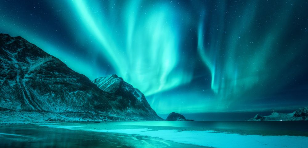Aurora Borealis lights up the night sky over snowy mountains and a frozen lake, casting a serene and magical glow in vibrant green and blue hues.