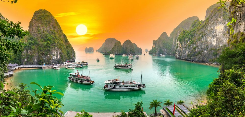 Sunset view of Halong Bay with emerald waters and limestone islands. Several boats are floating, creating a serene and picturesque seascape.