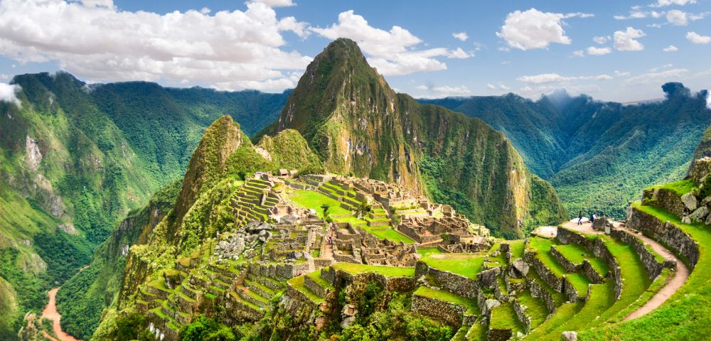 Scenic view of Machu Picchu with terraced ruins nestled in lush green mountains under a blue sky with fluffy clouds, conveying a sense of wonder and awe.