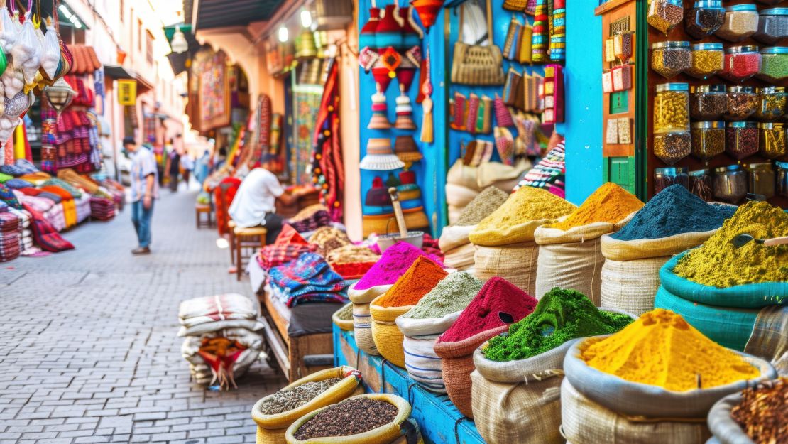 Vibrant market scene with sacks of colorful spices, including red, yellow, and green, against a backdrop of jars and woven goods.