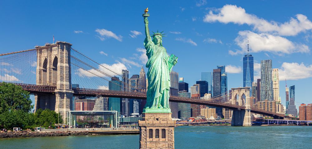 Statue of Liberty in the foreground, with the Brooklyn Bridge and Manhattan skyline under a bright blue sky. The scene conveys iconic New York City energy.