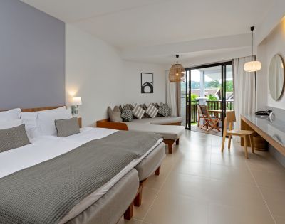 A modern hotel room features two beds with gray blankets, a cozy seating area, and a balcony view. The tone is serene and inviting, with soft lighting.