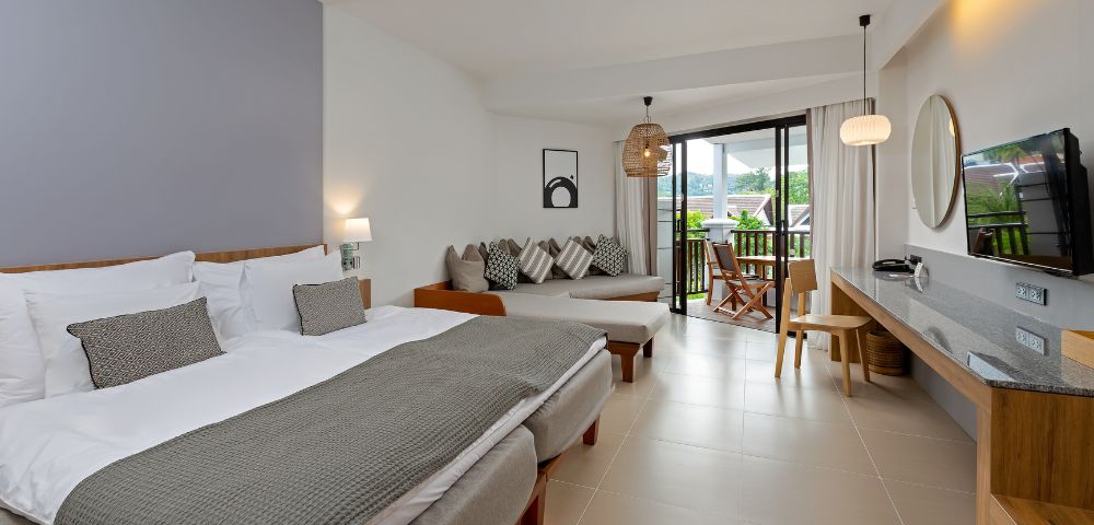 A modern hotel room features two beds with gray blankets, a cozy seating area, and a balcony view. The tone is serene and inviting, with soft lighting.