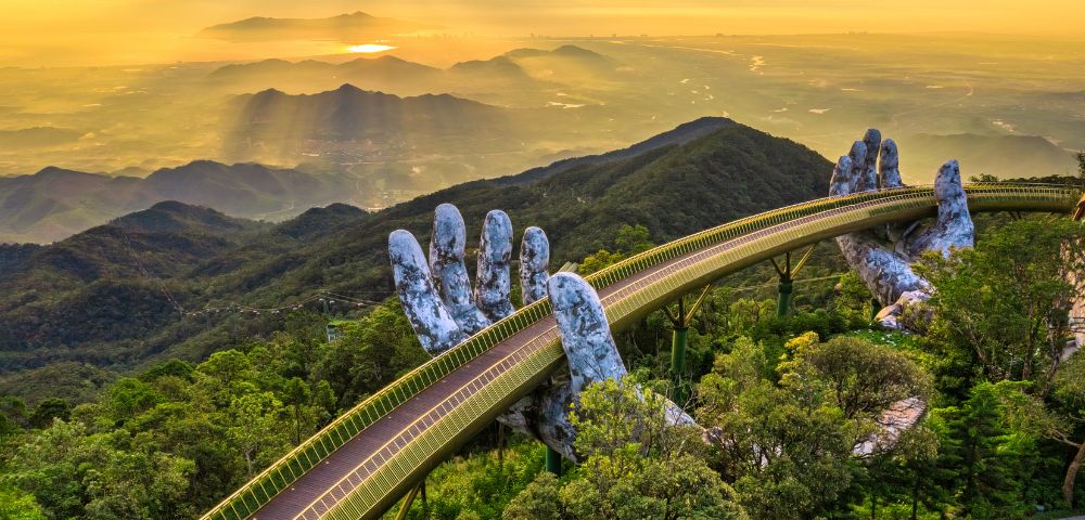 Golden bridge with giant stone hands emerges from lush forest, set against a glowing sunset over distant mountains and valleys, conveying awe and serenity.